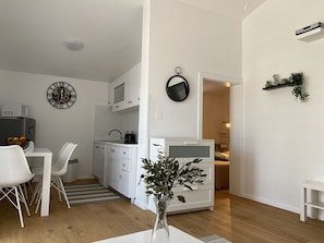 Une cuisine compacte avec des meubles blancs, un coin repas avec des chaises blanches, une petite table et une plante en pot.