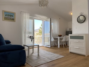 Un salon moderne avec un canapé bleu, une petite table sur laquelle est posé un vase, un meuble blanc et un balcon offrant une vue sur la campagne.