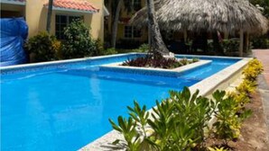 Una piscina con una estructura de techo de paja, rodeada de palmeras y un edificio con techo de tejas.