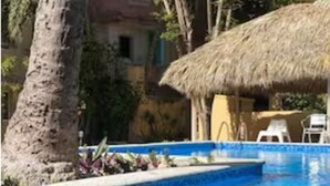 Una piscina con techo de paja, rodeada de árboles y un edificio de aspecto rústico.