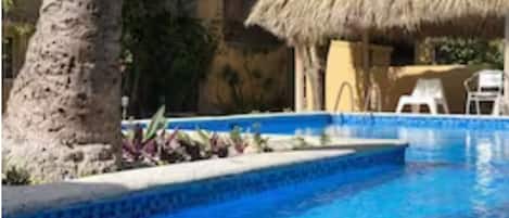 A swimming pool with a thatched roof structure, surrounded by trees and a building with a rustic appearance.