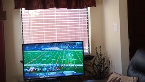 A living room with a television displaying a football game, a window with blinds, a decorative curtain, a leather sofa, and a floral rug.