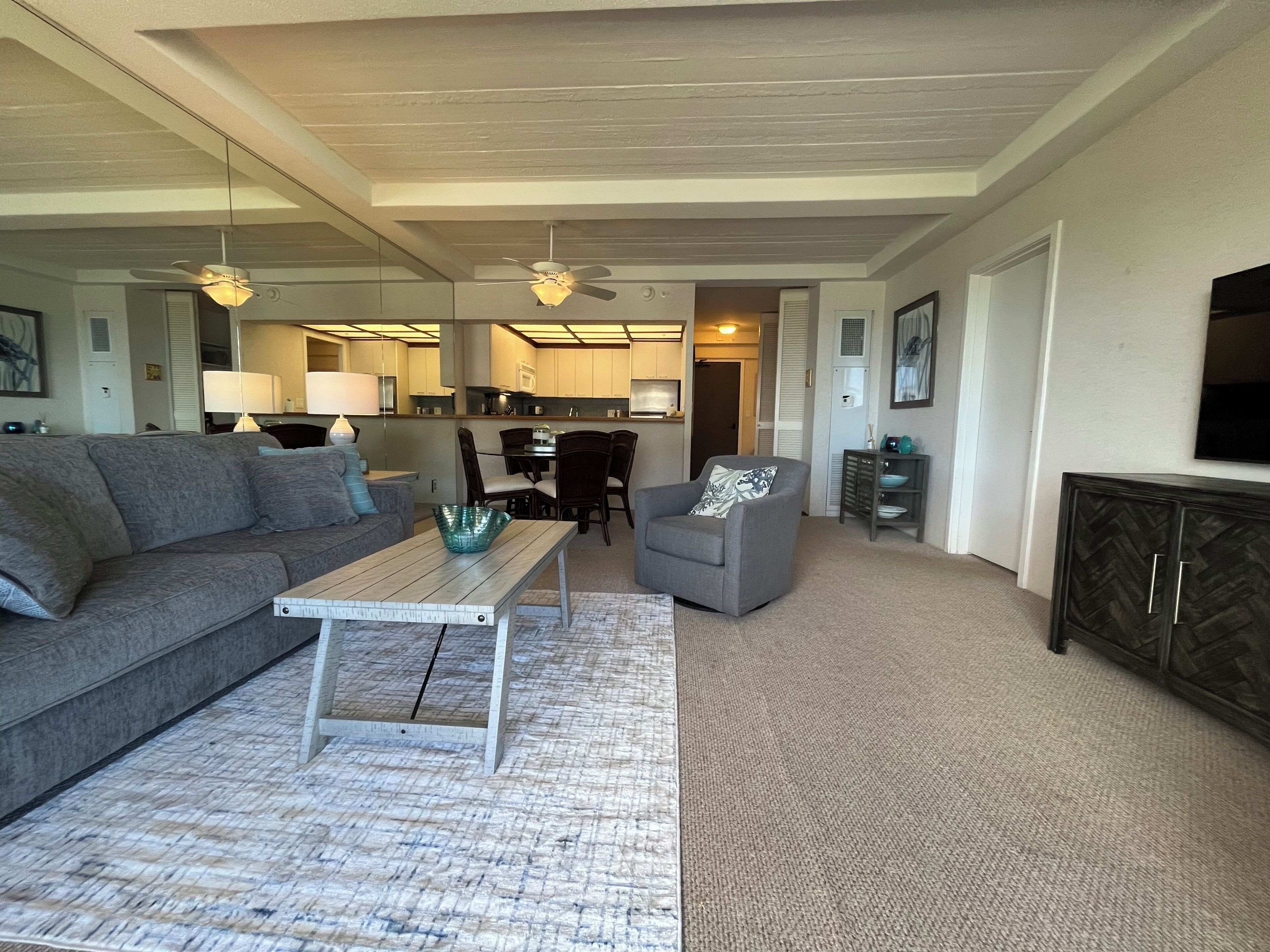 Ein modernes Wohnzimmer mit einem grauen Sofa, einem Couchtisch und einem grauen Sessel. Im Hintergrund ist eine Küchenzeile mit Esstisch und Stühlen zu sehen.