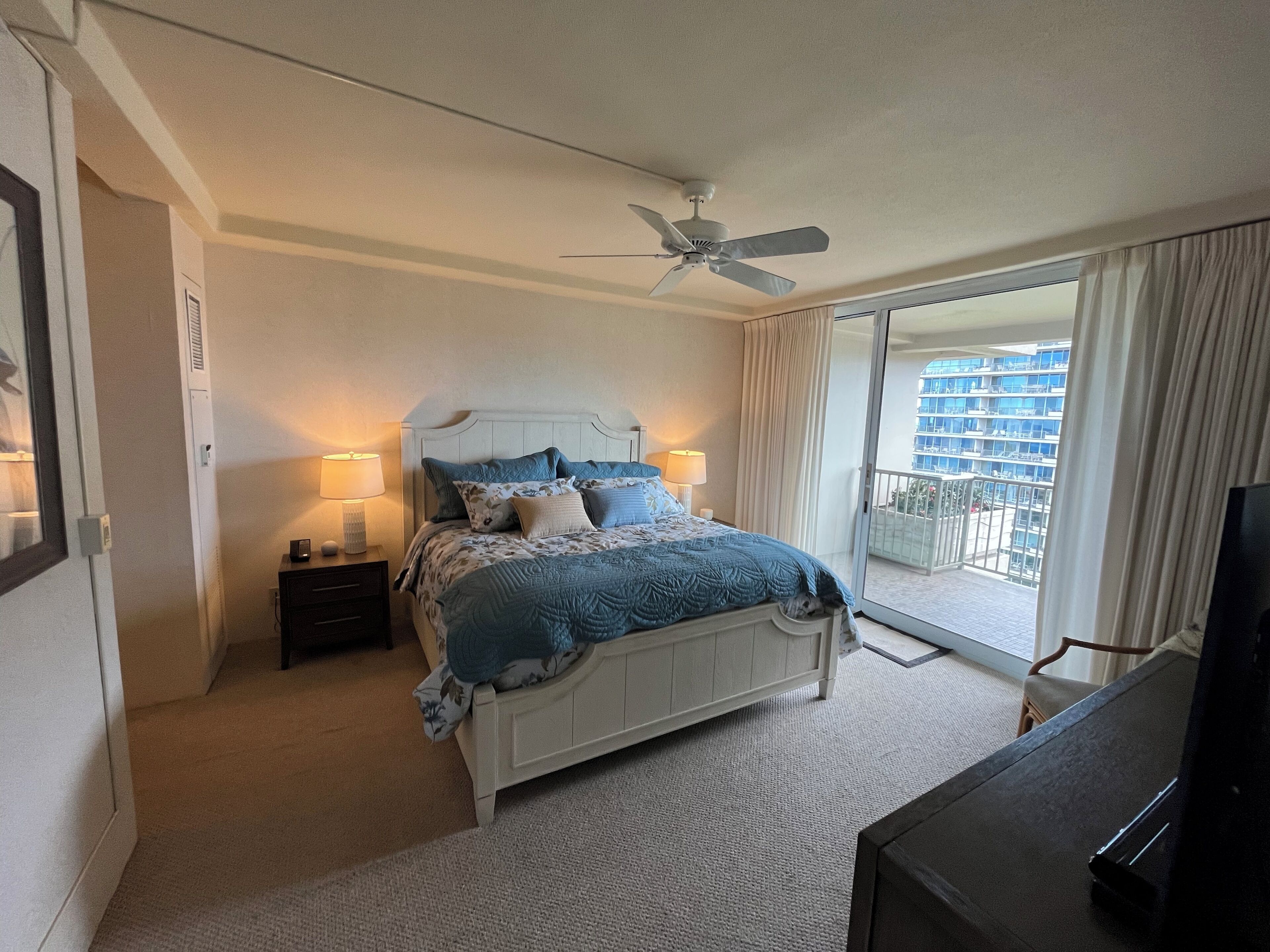 Hotelkamer met een bed, een plafondventilator, een nachtkastje met een lamp, een televisie en een balkon met uitzicht op gebouwen.