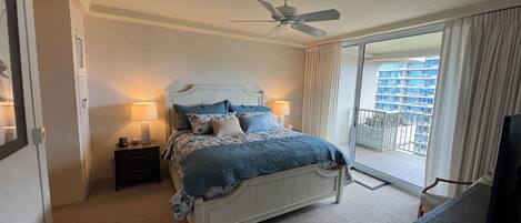 Hotelkamer met een bed, een plafondventilator, een nachtkastje met een lamp, een televisie en een balkon met uitzicht op gebouwen.