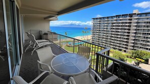 A balcony with a glass table and chairs, overlooking a beach and a hotel building. - Whaler Tower II - 1 Bedroom Condo with Beautiful Ocean View (Lahaina)