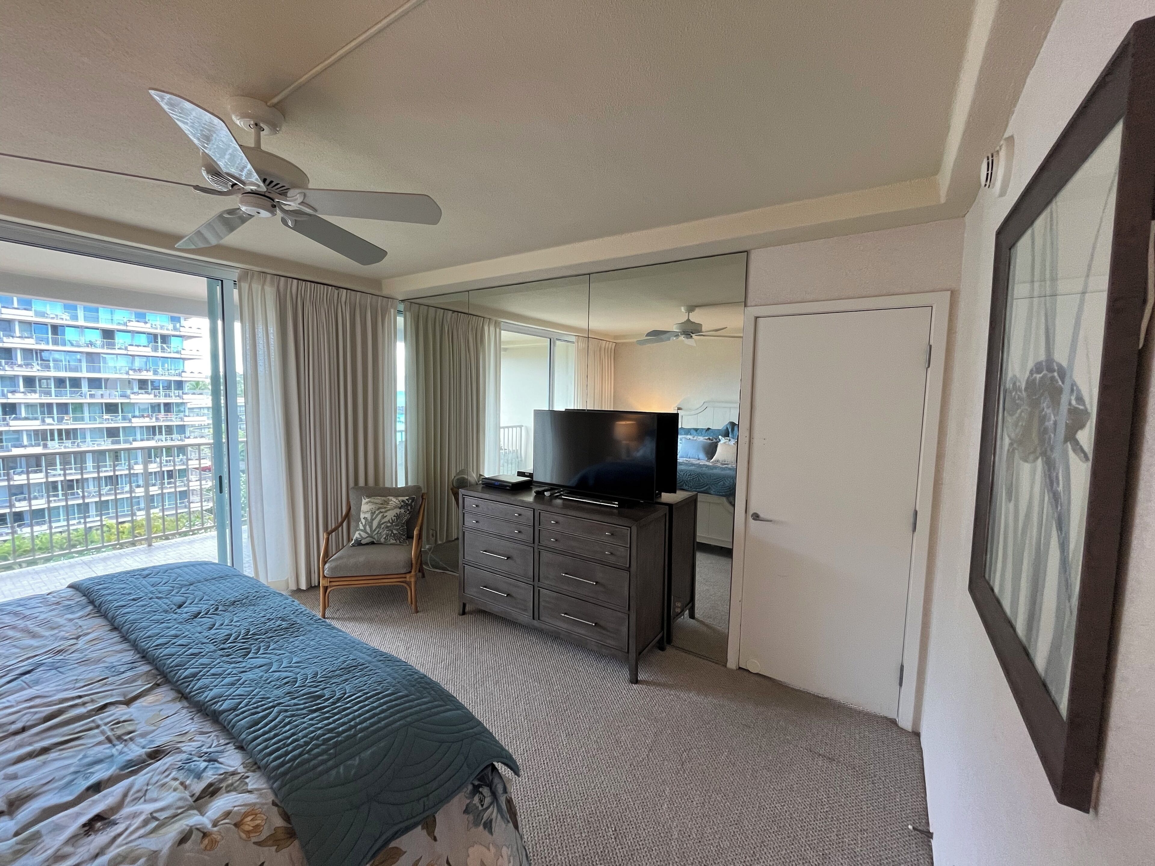 Ein Hotelzimmer mit einem Bett, einem Schminktisch, einem Fernseher, einem Deckenventilator und einem großen Fenster mit Vorhängen.