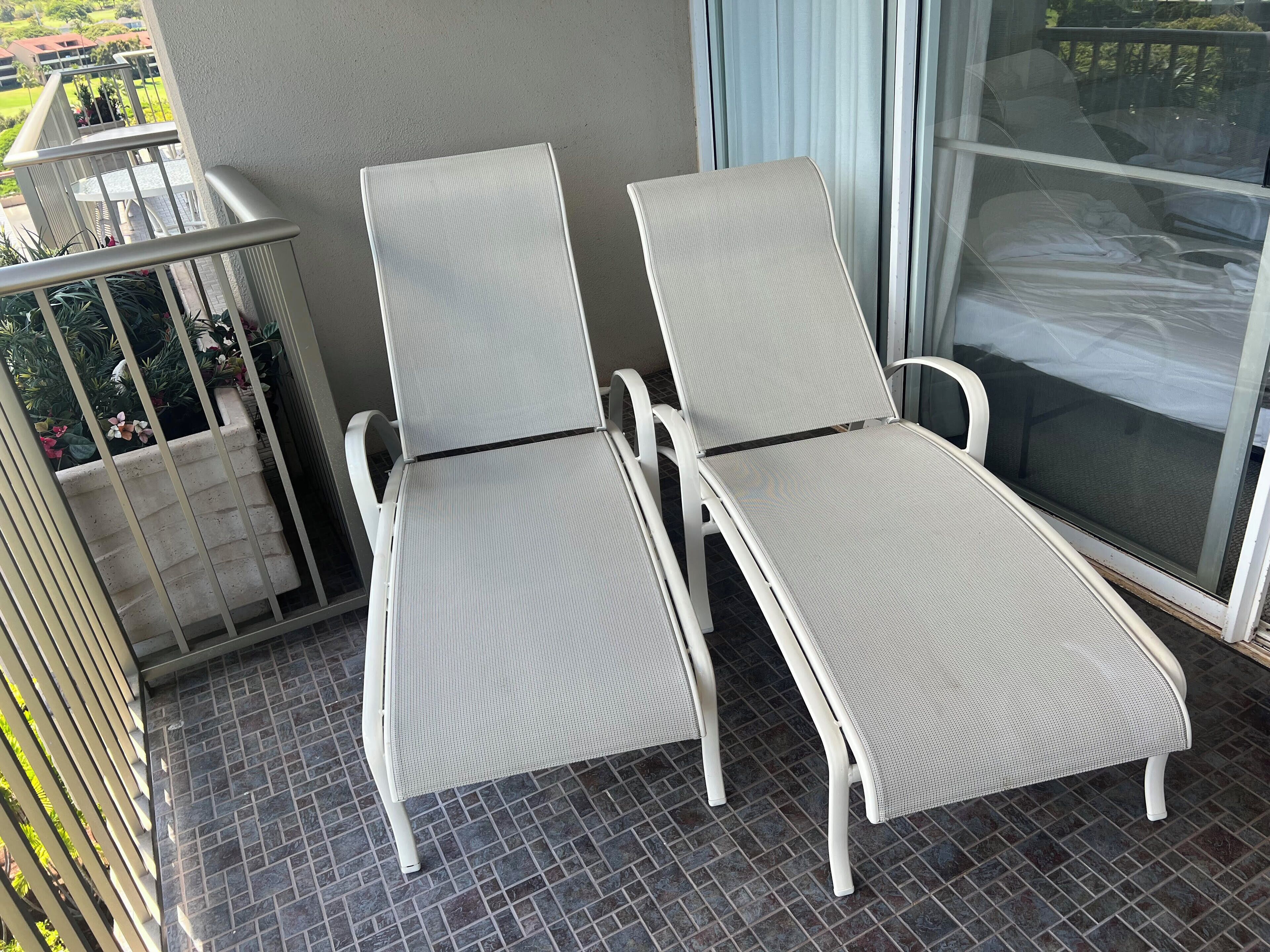 Two white lounge chairs with mesh backs and seats, positioned on a balcony with a view of a garden and a building in the background.