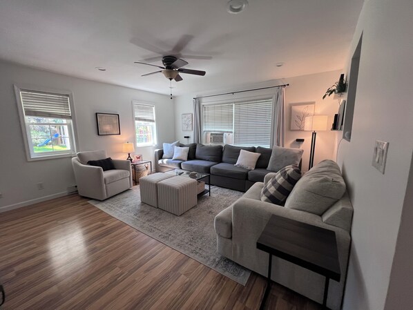 A spacious living room with a grey sectional sofa, a white armchair, and a glass coffee table. There is a ceiling fan, large windows with blinds, and a view of a playground outside.