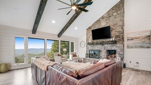 A spacious living room with a large leather sofa, a stone fireplace, and a television mounted on the wall. - Mountain-view home with high-speed WiFi, wood fireplace, W/D & central AC (Beech Mountain)