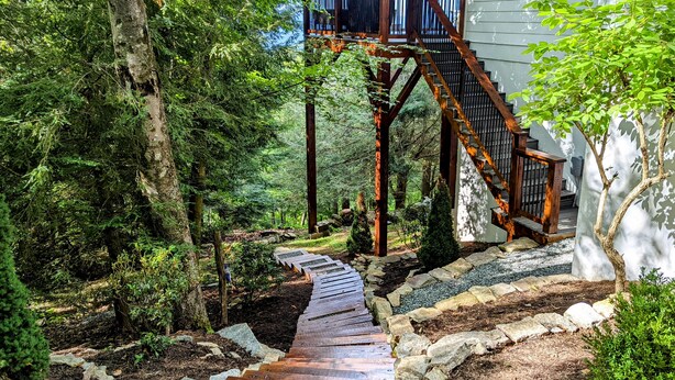 A wooden pathway leading to a wooden staircase with a metal railing, surrounded by trees and shrubs. - Mountain-view home with high-speed WiFi, wood fireplace, W/D & central AC (Beech Mountain)