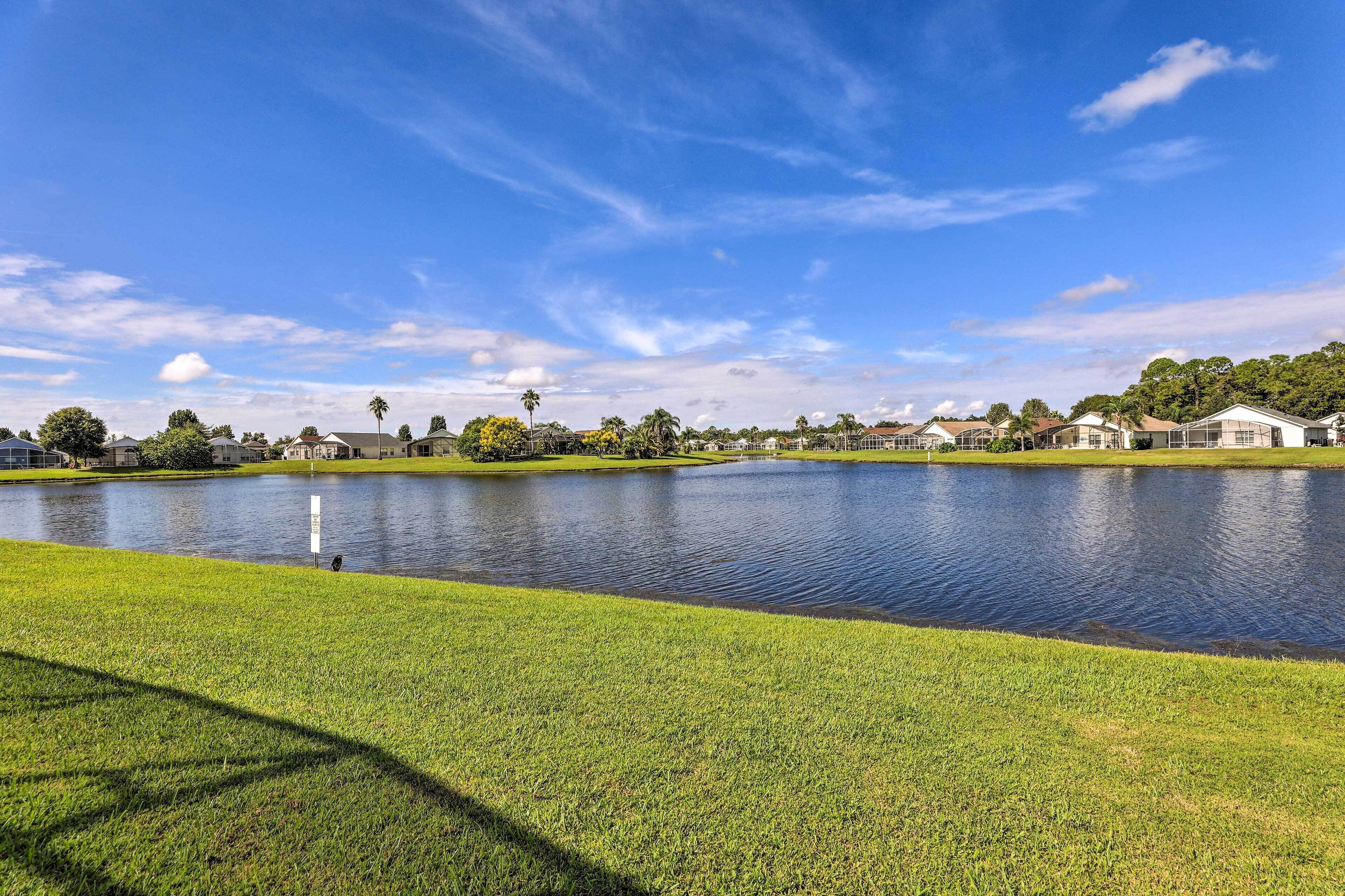 A serene lakeside view with a well-maintained grassy area, a calm lake, and a row of houses in the background.