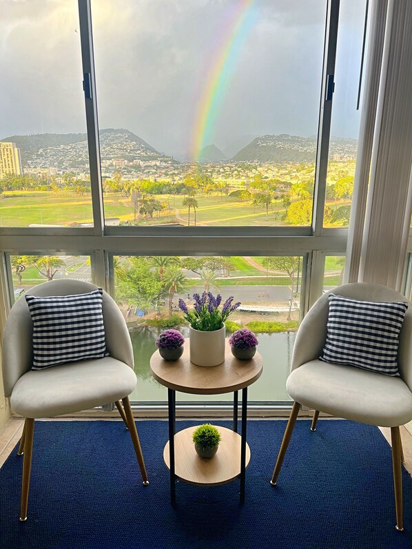 Une pièce avec deux fauteuils, une petite table ronde ornée d’un pot de fleurs, et une vue sur un arc-en-ciel se déployant au-dessus d’une ville.