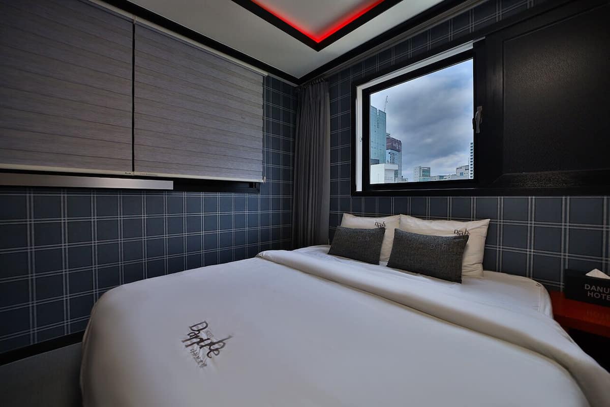 A modern hotel room with a large bed, a window with a city view, and a red accent on the ceiling.