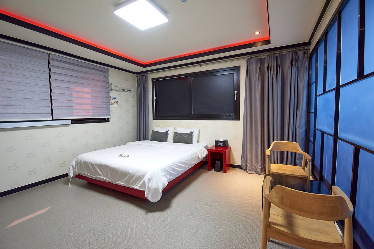 A hotel room with a bed, a small table, a chair, a window with curtains, and a red nightstand.