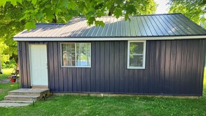 A small, single-story building with a dark exterior and a metal roof. It has a white door and two windows.