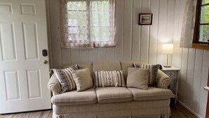 A beige sofa with patterned cushions, a white door, a window with lace curtains, a small table with a lamp, and a framed picture on the wall.