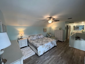 Une chambre à coucher comprenant un lit, des tables de chevet, une commode, un ventilateur de plafond et une salle de bain avec un miroir et un lavabo.
