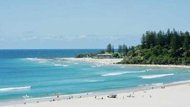A beach with clear turquoise waters, a sandy shore, and a line of trees in the background.