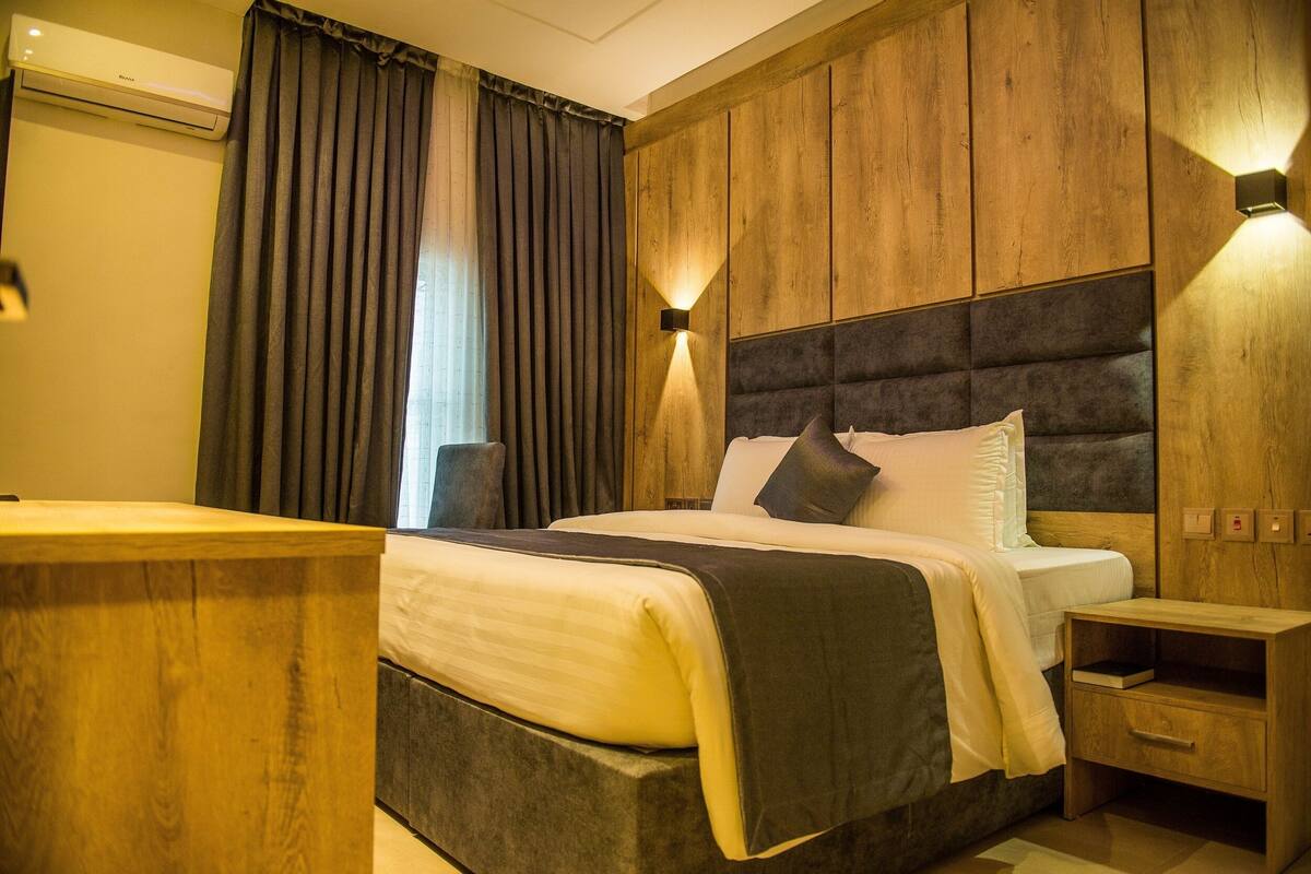 A hotel room with a large bed, wooden panel walls, a nightstand, and a wall-mounted light.