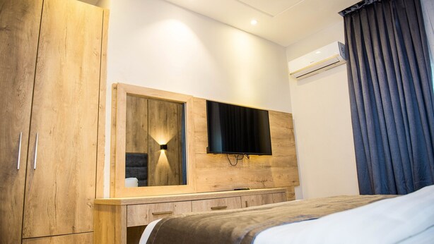 A bedroom with a wooden wardrobe, a bed, a flat-screen TV mounted on a wooden wall, and an air conditioning unit. - Pinnatech Suites (Abuja)