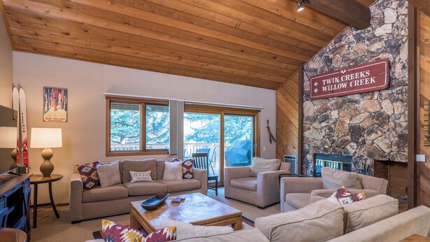 A cozy living room with a stone wall, wooden ceiling, and large windows. There is a sign indicating directions to Twin Creeks and Willow Creek. - Cozy Family sized Condo- access to Pools and other Summer time Amenities (Sun Valley)