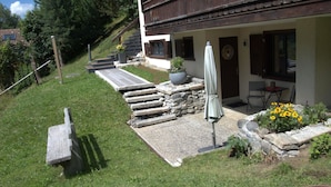 A house with a stone staircase leading to a wooden deck, a covered outdoor seating area, and a garden with yellow flowers. - Am Sonnenhang by Interhome (Churwalden)