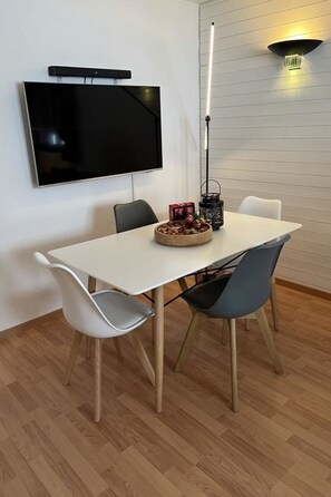 Comedor con una mesa blanca, dos sillas, un televisor de pantalla plana montado en la pared y una lámpara colgante moderna.