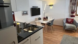 A compact kitchen with a dining area, a flat-screen TV, and a cozy seating area with a patterned throw. - Am Sonnenhang by Interhome (Churwalden)