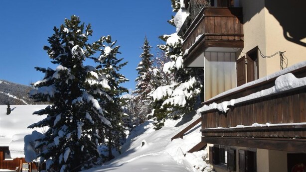 A snow-covered building with a wooden facade and a stone base, surrounded by snow-laden trees and a clear blue sky. - Am Sonnenhang by Interhome (Churwalden)