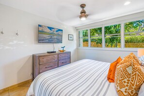 A bedroom with a bed, a dresser, a ceiling fan, a window with a view of greenery, and a painting on the wall.