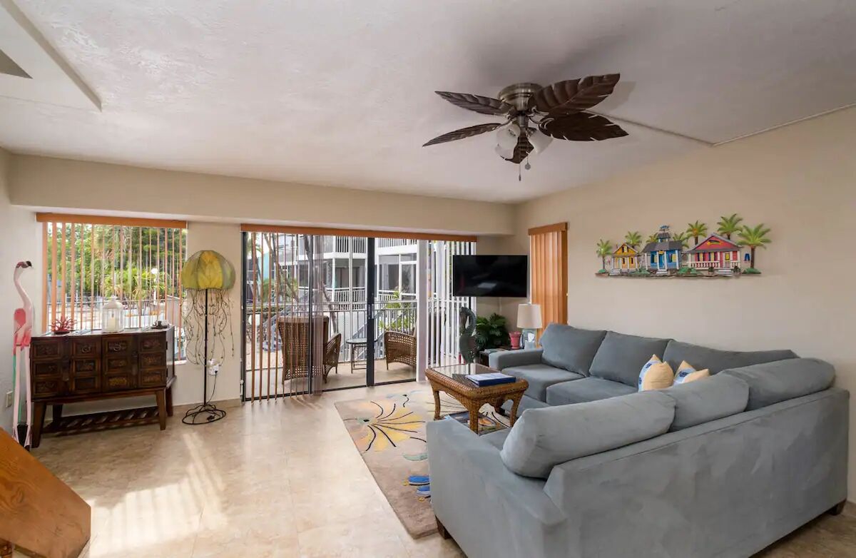 A living room with a grey sofa, a ceiling fan, a TV, and a window with a view of greenery.