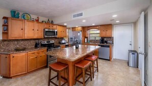 A kitchen with wooden cabinets, granite countertops, a central island with stools, stainless steel appliances, and a window with bars. - Free Golf Cart Free Kayaks Steps to Pool Pickleball (Key Largo)