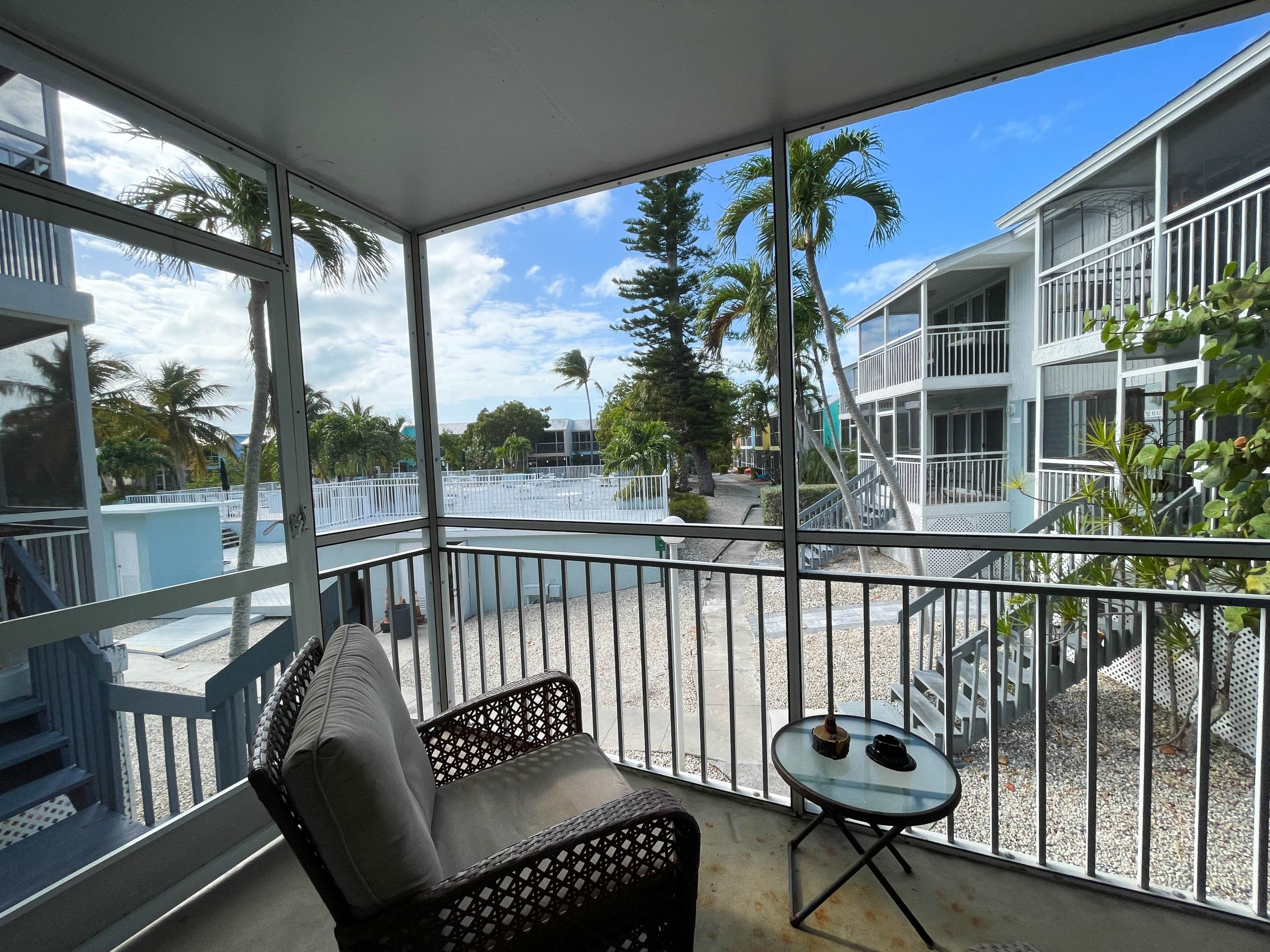 A balcony with a wicker chair and a small table, overlooking a building with balconies and palm trees.