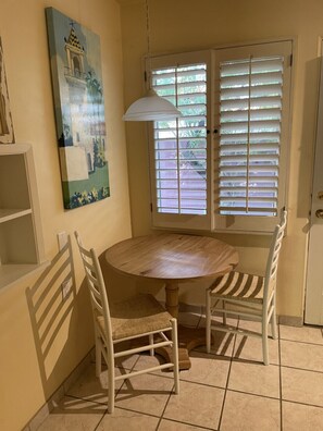A dining area with a round wooden table, two white chairs, a hanging pendant light, and a window with blinds. - Room 5️⃣ studio in a boutique hotel with kitchen by the pool in downtown PS  (Palm Springs)