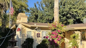 A building with a yellow facade, a flag, a fountain, and a garden with pink flowers.