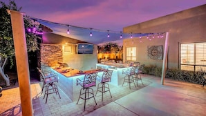An outdoor kitchen area with a bar and seating, surrounded by a pergola with string lights. - THE MASTERPIECE- Half Acre-8BR-5.5BA (Indio)