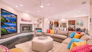 A spacious living room with a large flat-screen TV, a fireplace, and a kitchen in the background. - THE MASTERPIECE- Half Acre-8BR-5.5BA (Indio)