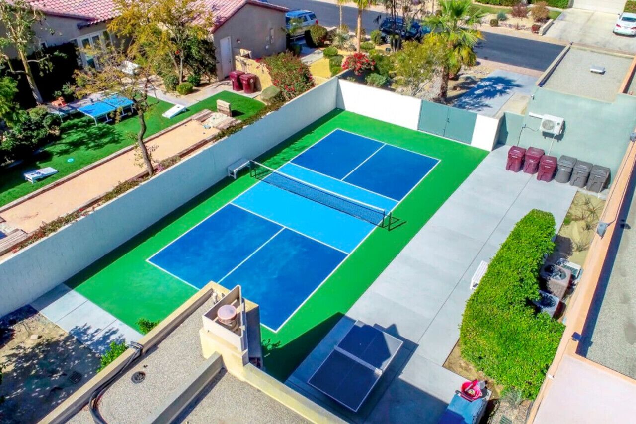 Un court de tennis extérieur doté d’une surface verte et bleue, entouré d’un petit muret et d’une aire de stationnement.