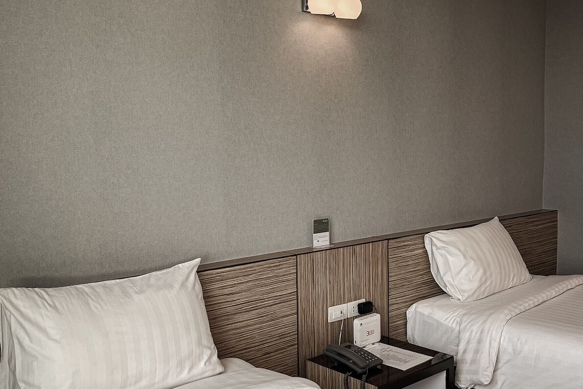 Habitación de hotel con dos camas, una mesita de noche con teléfono y lámpara, y una lámpara de pared.