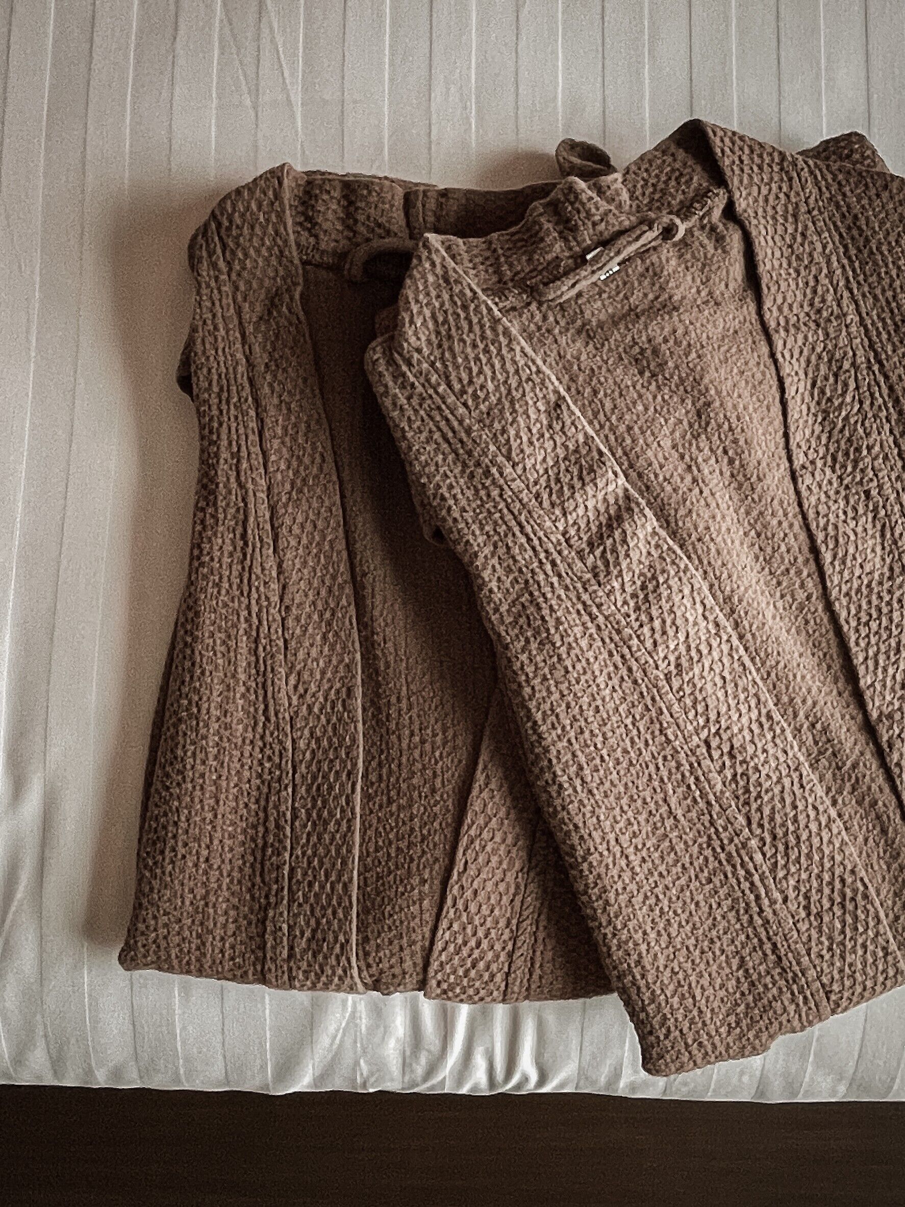 A bed with a white sheet and a brown knitted garment folded on it.