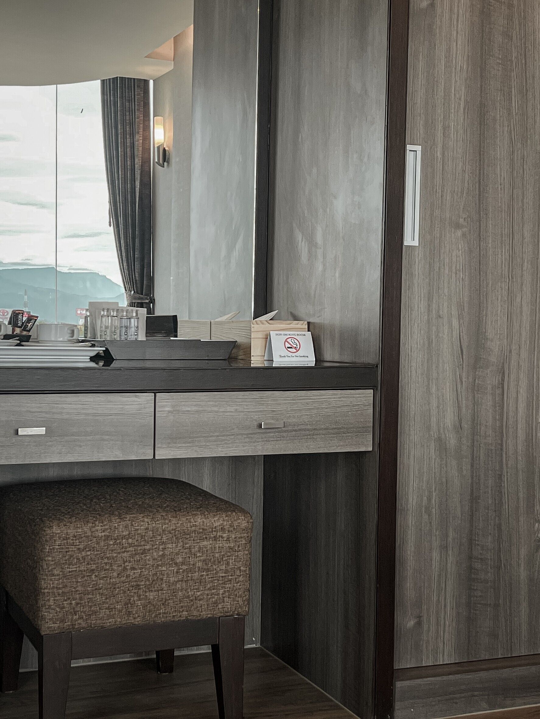 A modern hotel room with a large window, a dark wood panel wall, a small desk with a chair, and a sign indicating a non-smoking room.