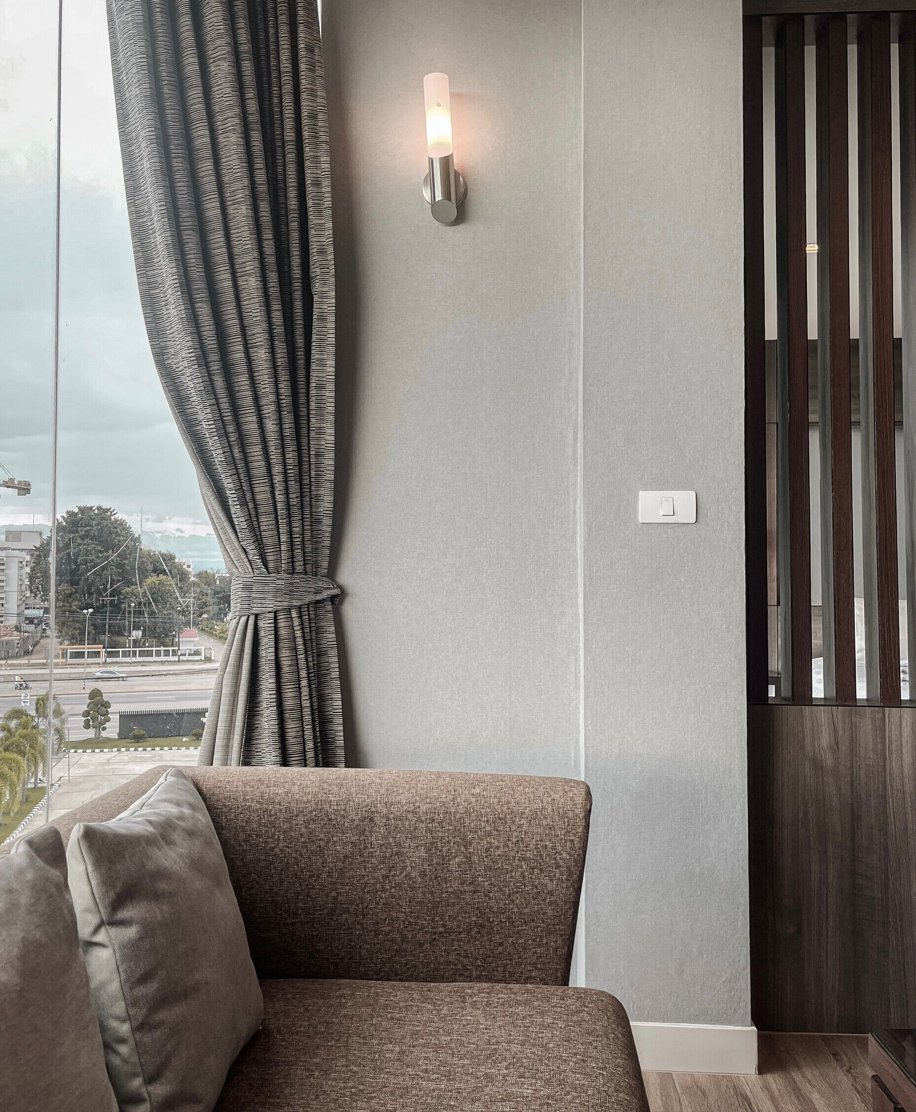 A modern living room with a grey sofa, a wall-mounted light fixture, and a view of a cityscape through a window with striped curtains.