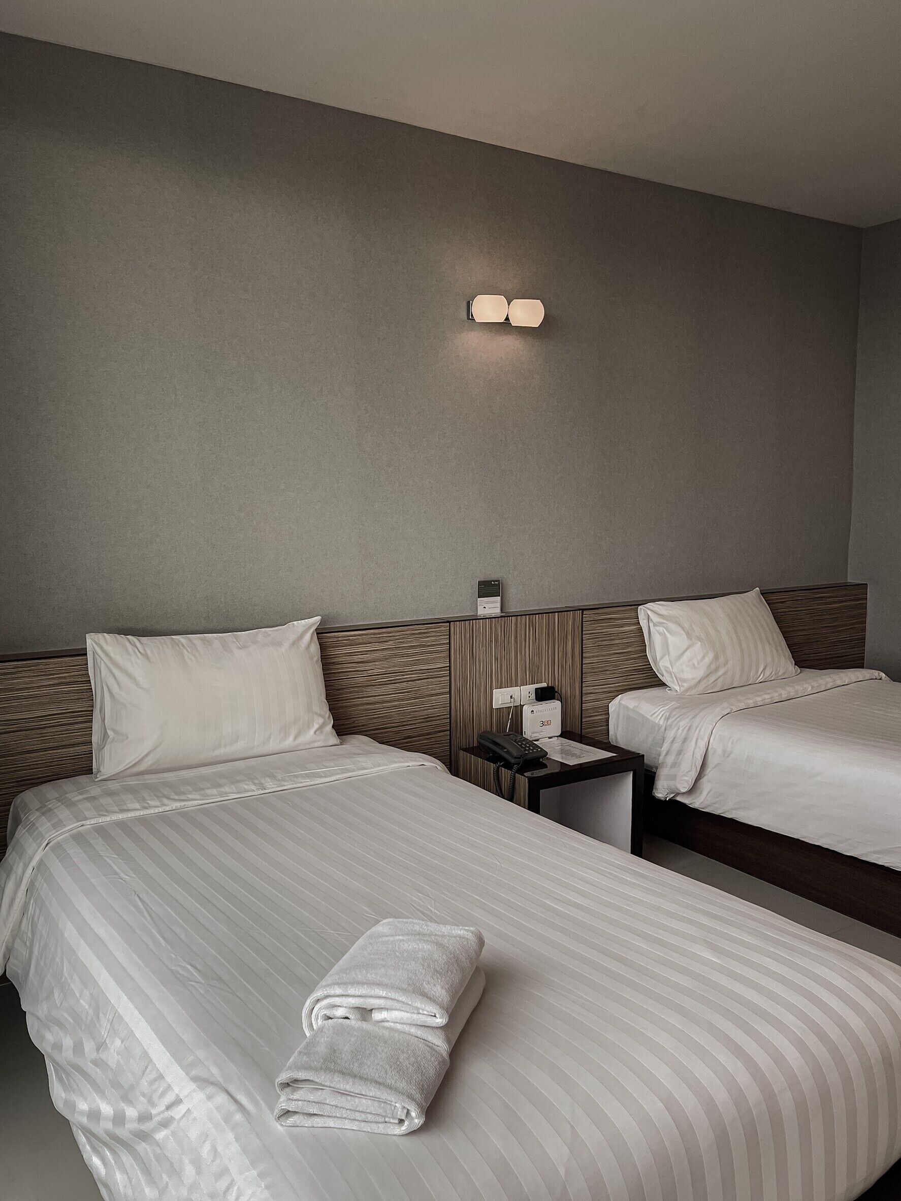 A hotel room with two beds, a nightstand with a phone, and a wall-mounted light fixture.