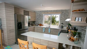 Een moderne keuken met een centraal eiland, houten stoelen, een koelkast en een raam met uitzicht op groen.
