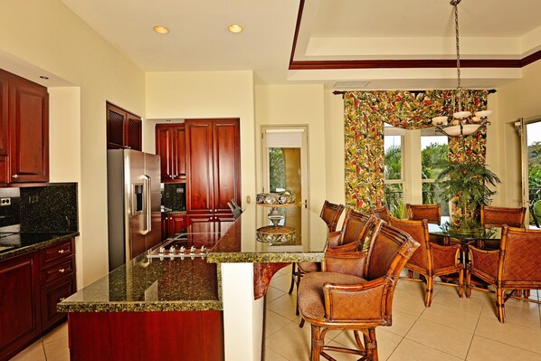 A kitchen with granite countertops, wooden cabinets, and a central island. There is a dining area with wicker chairs and a table.