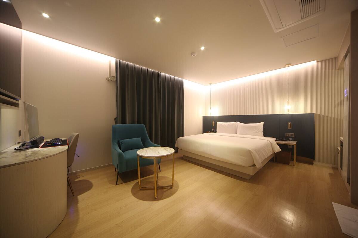 A modern hotel room with a bed, a desk with a computer, a chair, and a small table.