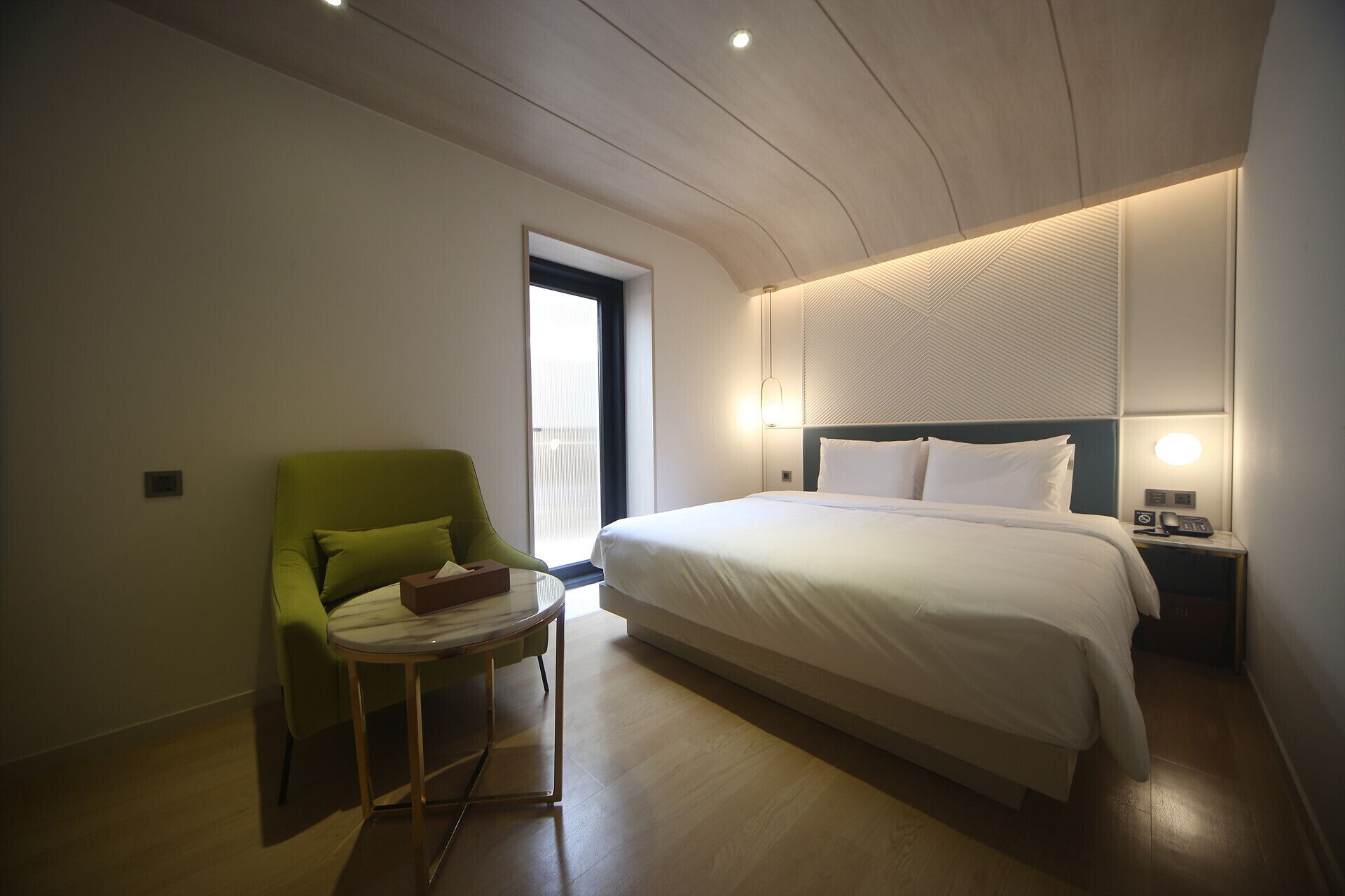 A modern hotel room with a large bed, a small table, a green armchair, and a nightstand with a lamp.