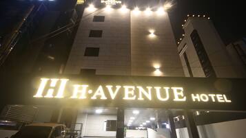 The exterior of the HI·AVENUE HOTEL with illuminated signage, a parking area, and a building facade.