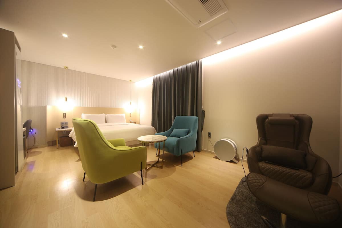 A hotel room with a bed, a green armchair, a blue armchair, a massage chair, and a small table.